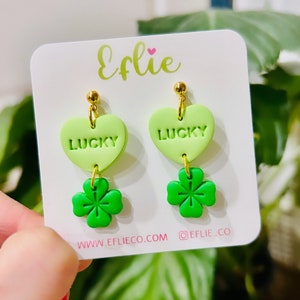 May include: A pair of green and light green heart-shaped earrings with the word "LUCKY" on each heart. Each heart is attached to a green four-leaf clover charm.