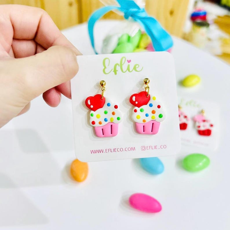 May include: A pair of cupcake-shaped earrings with a red heart on top. The cupcakes are white with colorful sprinkles and a pink base. The earrings are on a white card with the text "Efliie" and the website address "www.eflieco.com".