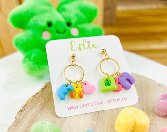 May include: Gold hoop earrings with colorful charms, including a heart, a four-leaf clover, a crescent moon, and a bear. The earrings are displayed on a white card with the text "Elie" and "www.eflieco.com @eflie_co".