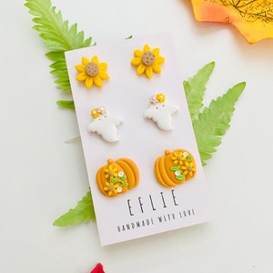 May include: A set of six polymer clay earrings featuring a pumpkin with yellow flowers, a ghost with a flower crown, and a sunflower. The earrings are displayed on a white card with the text "EFLIE HANDMADE WITH LOVE".