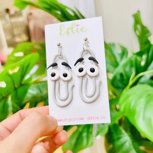 K&ouml;nnte beinhalten: Graue B&uuml;roklammer-Ohrringe mit wei&szlig;en Augen und schwarzen Augenbrauen. Die Ohrringe befinden sich auf einer wei&szlig;en Karte mit dem Text "Efiie" und "www.eflieco.com @eflie_co".