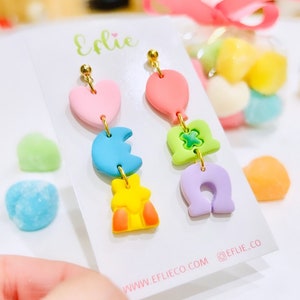 May include: Colorful polymer clay earrings featuring a heart, moon, star, balloon, four-leaf clover, and horseshoe charms. The earrings are in pastel shades of pink, blue, yellow, green, and purple, with gold-colored hardware. The card reads "Eflie" and "EFLIE.CO".