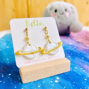 Op de afbeelding: Een paar witte planeetvormige oorbellen met gouden ringen en kleine gouden sterbedels bevestigd aan gouden stekers. De oorbellen worden gepresenteerd op een witte kaart met de tekst Eflie en www.eflieco.com, geplaatst op een licht houten blokje.