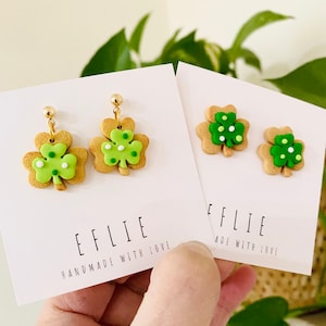 May include: A pair of green and brown shamrock-shaped earrings with white polka dots. The earrings are handmade and have gold hooks. The earrings are displayed on a white card with the text "EFLIE HANDMADE WITH LOVE".