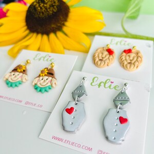 May include: Four pairs of polymer clay earrings inspired by the Wizard of Oz. The first pair features a scarecrow with a straw hat and a green shirt. The second pair features a lion with a red bow. The third pair features a tin man with a silver body and a red heart. The fourth pair features a witch with a black hat and a green dress. The earrings are on white cards with the text "Elie" and "www.eflieco.com @eflie_co".