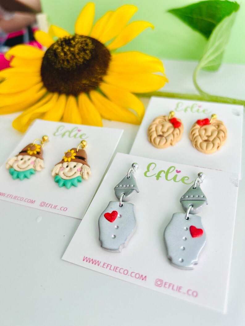 May include: Four pairs of polymer clay earrings inspired by the Wizard of Oz. The first pair features a scarecrow with a straw hat and a green shirt. The second pair features a lion with a red bow. The third pair features a tin man with a silver body and a red heart. The fourth pair features a witch with a black hat and a green dress. The earrings are on white cards with the text "Elie" and "www.eflieco.com @eflie_co".