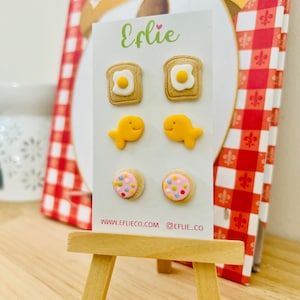 May include: A set of three pairs of novelty earrings displayed on a small easel. The earrings include pairs of toast with fried eggs, goldfish crackers, and pink cookies with colorful sprinkles. The brand name "Eflie" is printed in green.