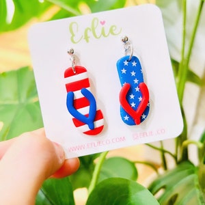 May include: A pair of red, white, and blue flip-flop earrings. One earring features a red, white, and blue striped flip-flop, while the other earring features a blue flip-flop with white stars. The earrings are on a white card with the text "Efflie" and "www.efflieco.com" printed on it.