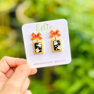 Pode incluir: Um par de brincos pendentes num cartão expositor branco com a marca Eflie. Cada brinco apresenta um laço laranja preso a um pendente retangular em forma de pastel com cobertura preta, figuras de fantasmas brancos e granulados coloridos.