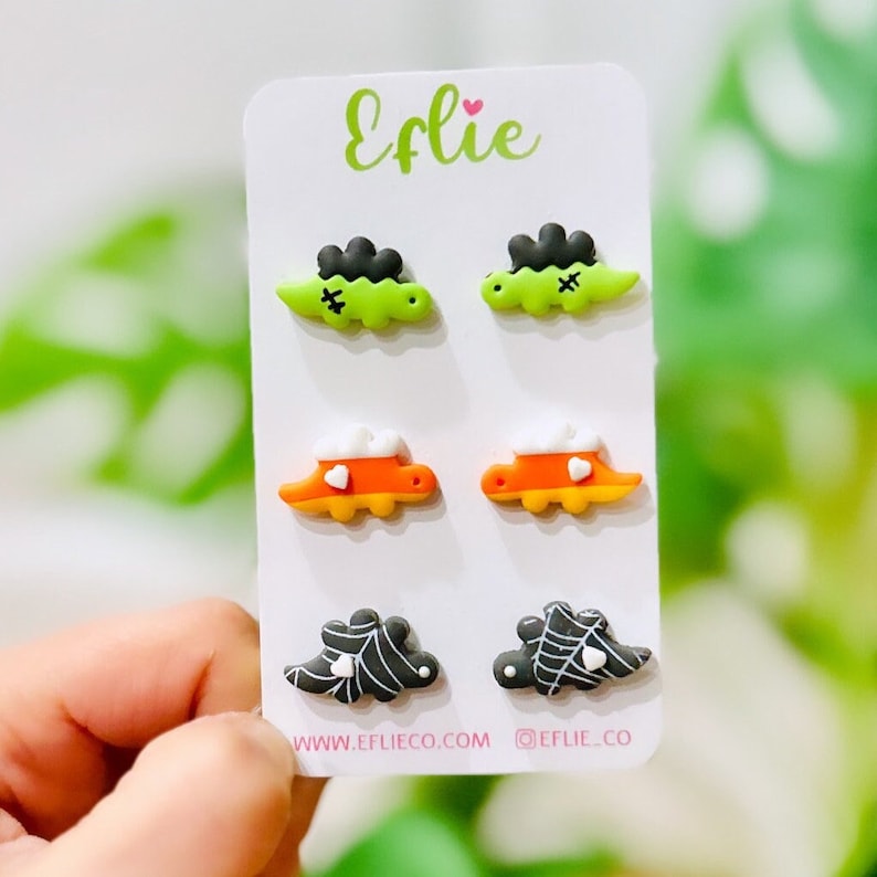 K&ouml;nnte beinhalten: Ein Set aus sechs Halloween-Ohrsteckern auf einer wei&szlig;en Karte. Die Ohrringe zeigen Designs von gr&uuml;nen Frankenstein-Monstern, orangefarbenen Zuckermais und schwarzen Spinnennetzen. Die Karte tr&auml;gt den Markennamen "Eflie" in gr&uuml;ner Schrift.