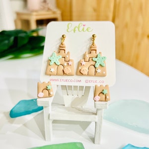 Puede incluir: Un par de pendientes de castillo de arena con una estrella de mar, una concha y estrellas blancas. Los pendientes se muestran en una tarjeta blanca con el texto "EFLIE" y "www.eflieco.com @eflie_co".