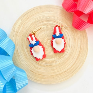 May include: A pair of red, white, and blue gnome earrings with gold hooks. The gnomes are wearing a red hat with a white stripe and a blue beard.