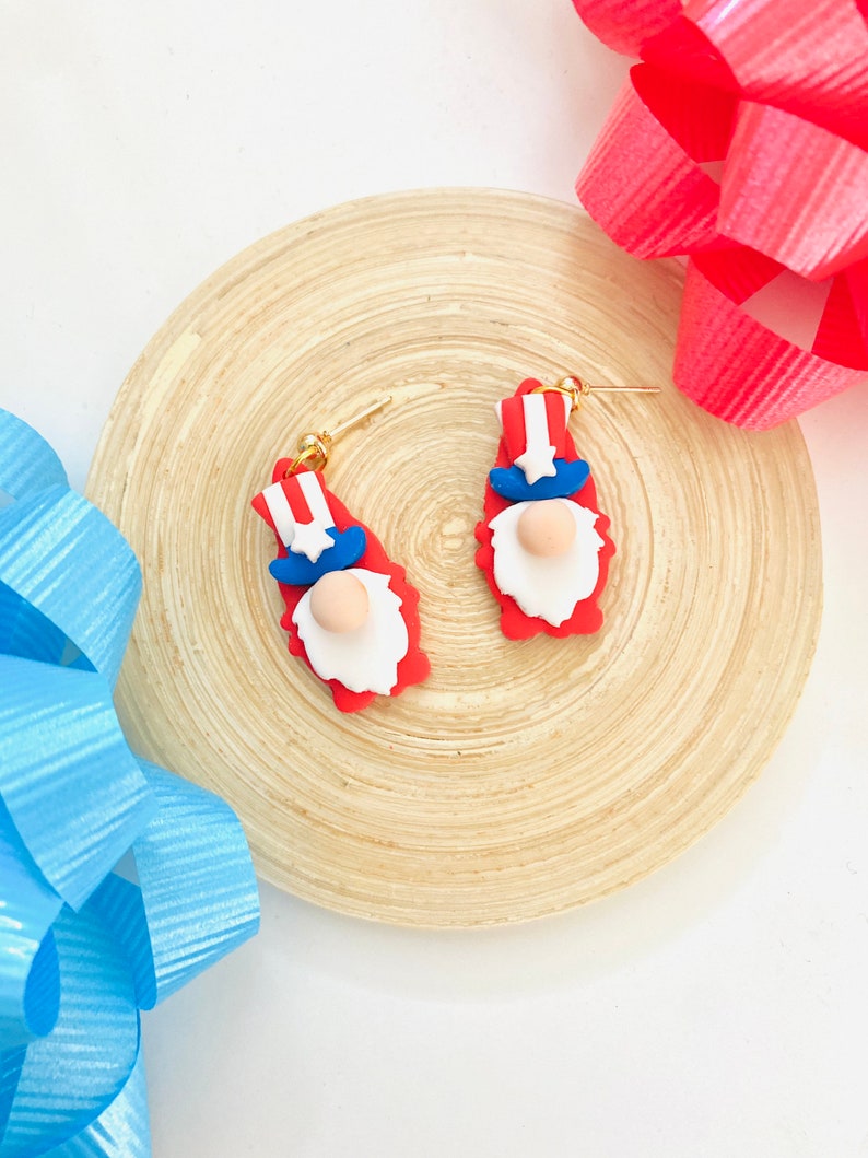 May include: A pair of red, white, and blue gnome earrings with gold hooks. The gnomes are wearing a red hat with a white stripe and a blue beard.