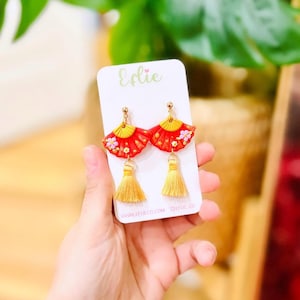 May include: Red fan-shaped earrings with yellow detailing and gold tassels. The earrings are decorated with small pink flowers and presented on a white card with the name "Eflie" in green script. The website address is visible.