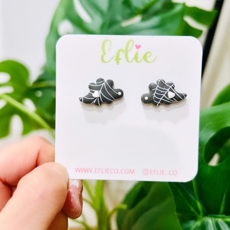 K&ouml;nnte beinhalten: Ein Paar schwarz-wei&szlig;e Halloween-Ohrstecker. Jeder Ohrring hat ein stilisiertes Design mit einem Spinnennetz und einem kleinen Herz. Die Ohrringe werden auf einer wei&szlig;en Karte mit dem Markennamen "Eflie" pr&auml;sentiert.