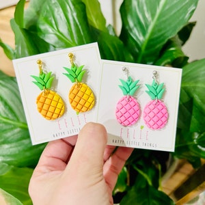 May include: Two pairs of pineapple earrings on white cards. The earrings are made of polymer clay and have gold or silver ear wires. The pineapples are yellow and orange or pink. The cards have the text "EFFLIE HAPPY LITTLE THINGS" on them.