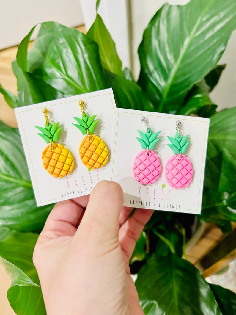 May include: Two pairs of pineapple earrings on white cards. The earrings are made of polymer clay and have gold or silver ear wires. The pineapples are yellow and orange or pink. The cards have the text "EFFLIE HAPPY LITTLE THINGS" on them.