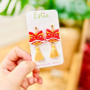 May include: A pair of red, yellow, and white lion dance earrings with gold-coloured tassels. The earrings are on a white card with the text "Eflie" and the website address www.eflieco.com.