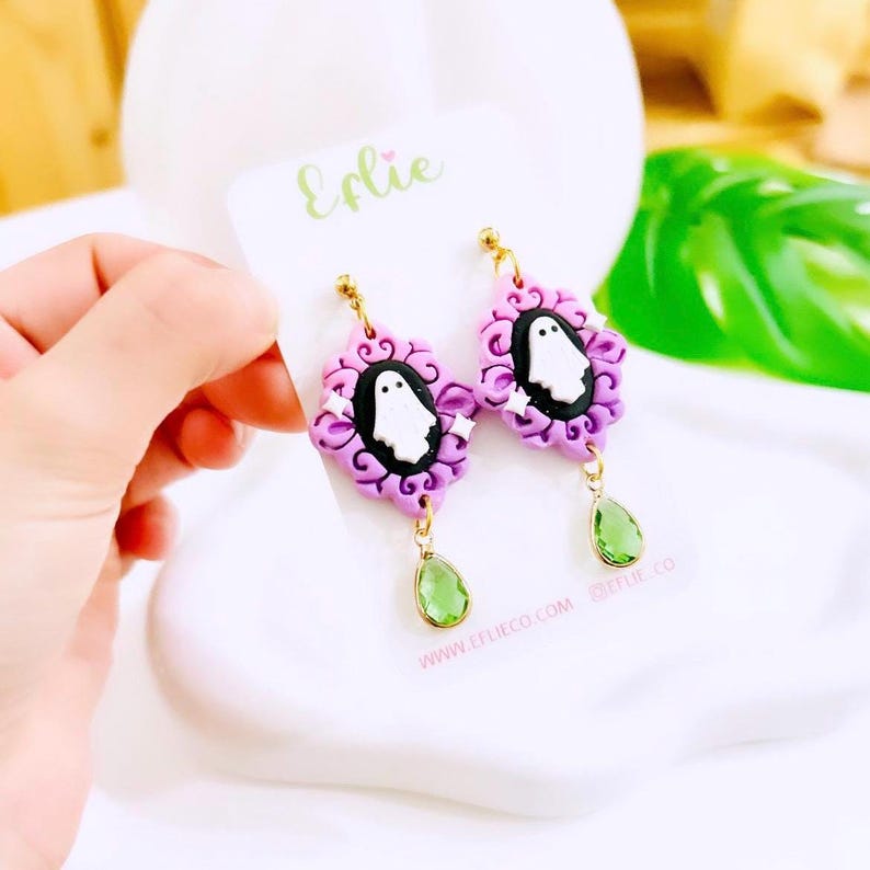 May include: A pair of handmade earrings featuring a ghost design. The earrings have a purple frame with a white ghost on a black background, accented with white stars. Each earring has a green teardrop charm. The brand name "Erlie" is visible.