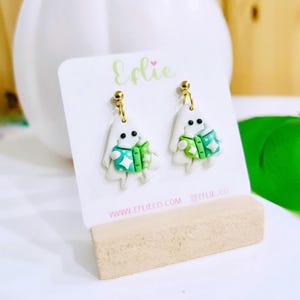 Op de afbeelding: Handgemaakte spookoorbellen. Elke witte spookfiguur heeft een groen en blauwgroen jasje met sterdetails en zwarte ogen. De oorbellen hangen aan goudkleurige stiftjes. De merknaam "Eflie" is zichtbaar op de kaart.