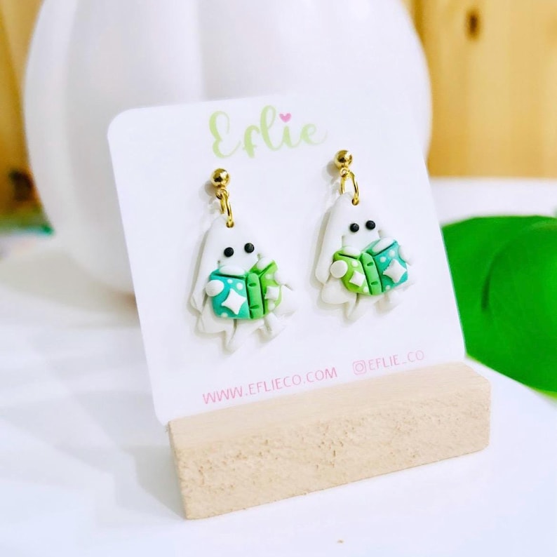 Op de afbeelding: Handgemaakte spookoorbellen. Elke witte spookfiguur heeft een groen en blauwgroen jasje met sterdetails en zwarte ogen. De oorbellen hangen aan goudkleurige stiftjes. De merknaam "Eflie" is zichtbaar op de kaart.