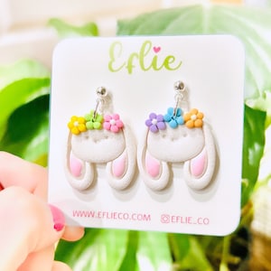 May include: A pair of white bunny-shaped earrings with pastel pink ears and a flower crown. The flowers are in shades of yellow, green, blue, and pink. The earrings are on a white card with the text "Efie" and "www.eflieco.com @eflie_co".