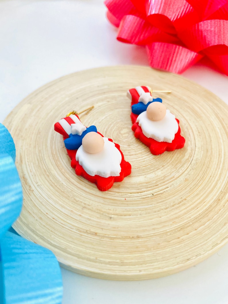 May include: A pair of red, white, and blue gnome earrings. The gnomes are wearing a blue hat with a white star and a white beard.
