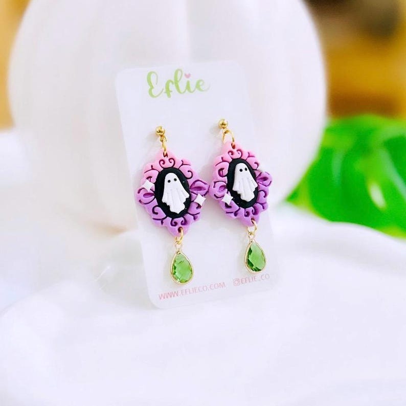 May include: A pair of Halloween-themed earrings. Each earring features a white ghost in a black oval frame, surrounded by pink and purple decorative elements. Green teardrop-shaped gems dangle below. The earrings are on a white card with the name "Erlie".