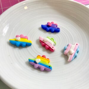 May include: Five colorful dinosaur charms with white hearts on a white plate. The dinosaurs are made of polymer clay and are painted in various colors, including blue, green, pink, yellow, and rainbow.