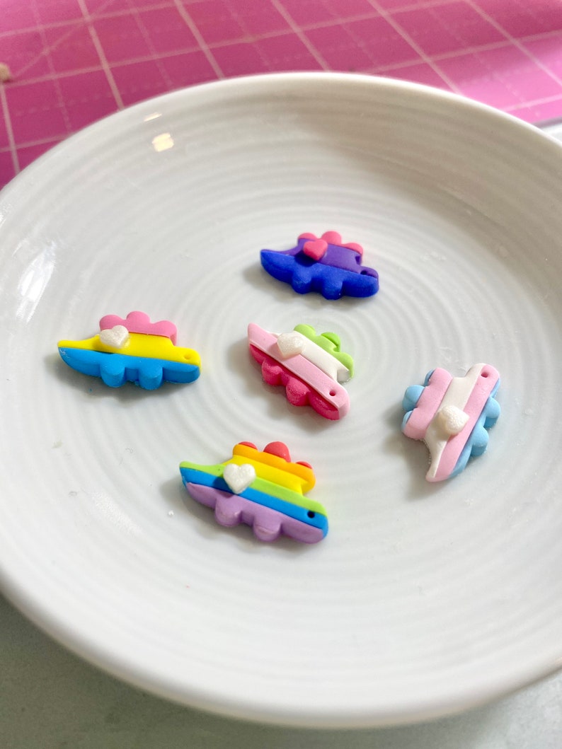 May include: Five colorful dinosaur charms with white hearts on a white plate. The dinosaurs are made of polymer clay and are painted in various colors, including blue, green, pink, yellow, and rainbow.
