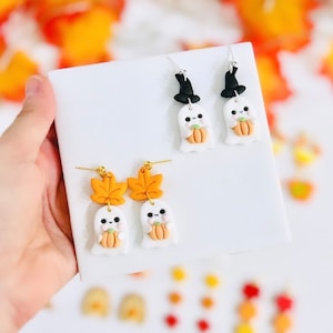 May include: A pair of dangle earrings featuring a white ghost holding a small orange pumpkin. One pair has a black witch hat on top of the ghost and the other pair has a small orange leaf charm.