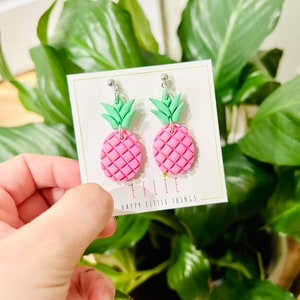 May include: Pink and green pineapple shaped earrings with silver hooks. The earrings are on a white card with the text "EFLIE HAPPY LITTLE THINGS".