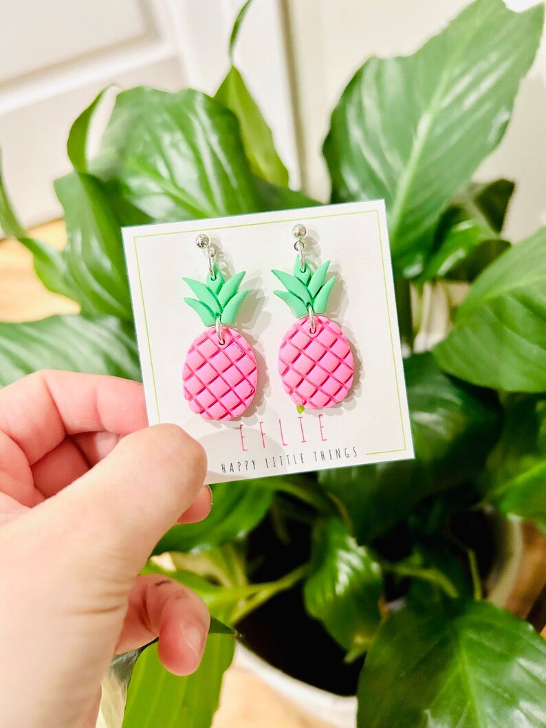May include: Pink and green pineapple shaped earrings with silver hooks. The earrings are on a white card with the text "EFLIE HAPPY LITTLE THINGS".