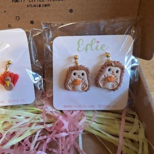 May include: A pair of brown and white hedgehog earrings with gold hooks. The earrings are on a white card with the word "Efie" written in green. The card also has the website address "www.eflieco.com" and the social media handle "@eflie.co".