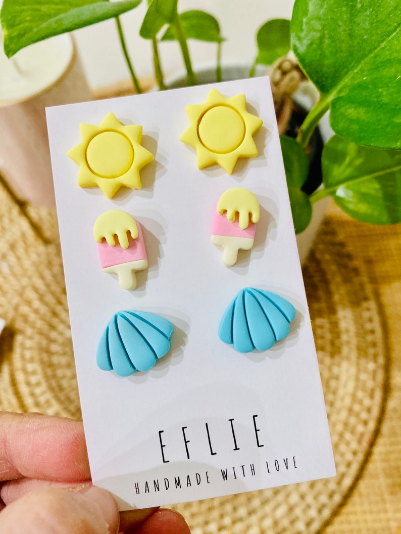 May include: A set of six colorful polymer clay earrings featuring a sun, ice cream, and seashell design. The earrings are displayed on a white card with the text "EFLIE HANDMADE WITH LOVE".