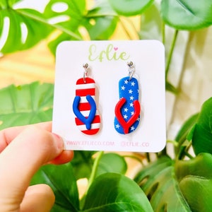 May include: A pair of red, white, and blue flip-flop earrings. The flip-flops are decorated with a red, white, and blue American flag pattern. The earrings are on a white card with the text "Efliie" and "www.efliieco.com @efliie_co".
