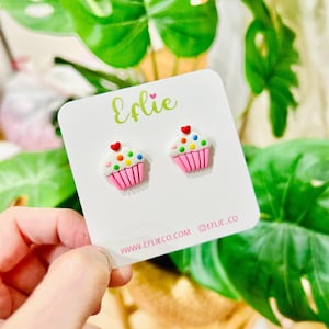 May include: Cupcake stud earrings on a white card. The earrings feature pink cupcake cases, white icing, and colourful sprinkles, topped with a red heart. The card displays the word "Eflie" in green script.