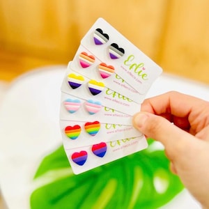Pode incluir: Uma coleção de brincos de coração em vários designs de bandeiras do orgulho. Cada par é montado em um cartão branco com o nome da marca "Erlie" e o endereço do site. Os brincos apresentam cores como arco-íris, rosa, azul e roxo.