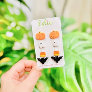 Puede incluir: Un conjunto de seis pendientes de botón con temática de Halloween. Los pendientes presentan un diseño de calabaza, fantasma, maíz dulce y murciélago. Los pendientes se muestran en una tarjeta blanca con el texto "Efliie" y "www.eflieco.com @eflie.co".