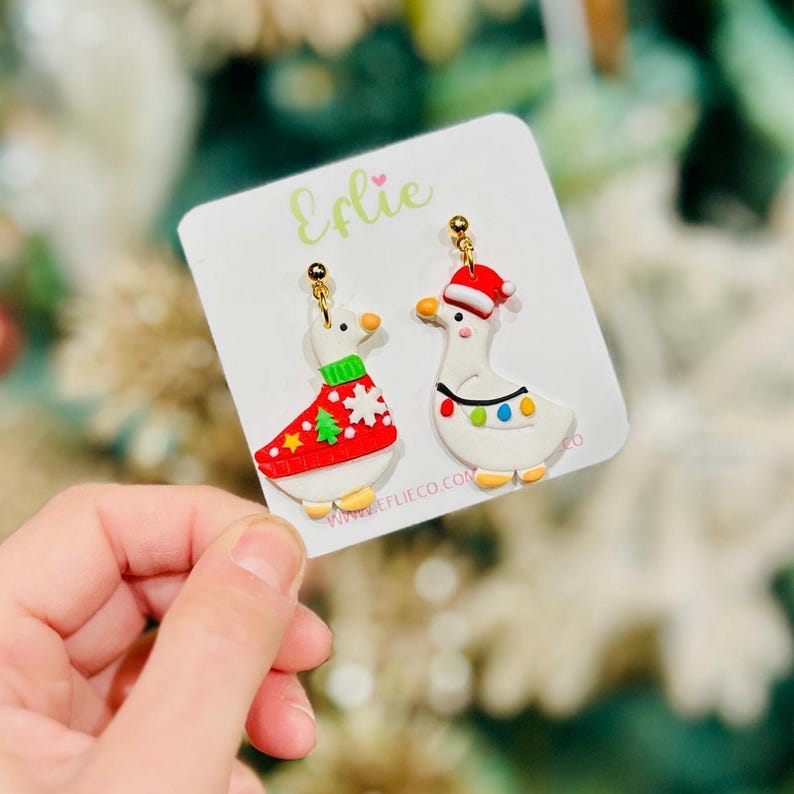 Op de afbeelding: Een paar kerstoorbellen met witte ganzenontwerpen. E&eacute;n gans draagt een rode trui met een kerstboom, de andere een kerstmuts en lichtslingers. De oorbellen zitten op een witte kaart met de naam "Eflie".