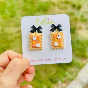 May include: A pair of Halloween-themed earrings. Each earring features a rectangular biscuit design with an orange center, white ghost figures, and a black bow. The earrings are held against a green background.