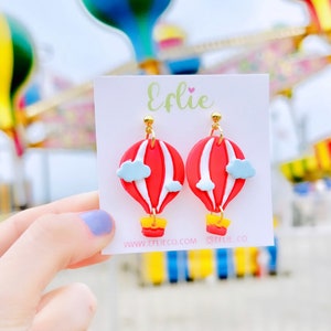 May include: A pair of red, white, and blue hot air balloon earrings with gold accents. The earrings are on a white card with the text "Efie" and "www.eflieco.com" and "@eflie.co".