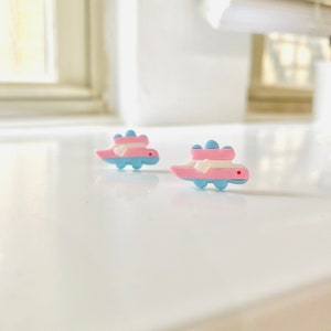 May include: Two small, blue and pink dinosaur-shaped earrings with white bows. The earrings are on a white surface.