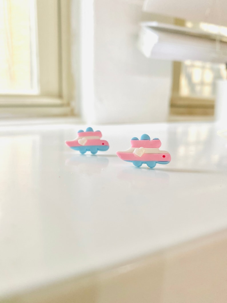 May include: Two small, blue and pink dinosaur-shaped earrings with white bows. The earrings are on a white surface.
