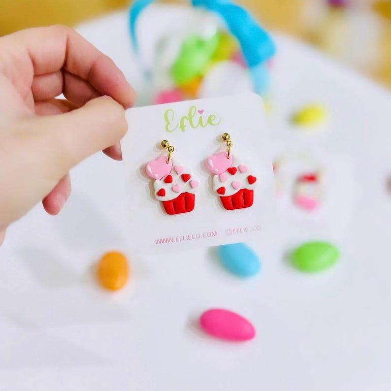 May include: A pair of red, white, and pink cupcake earrings with gold hooks. The cupcakes have pink hearts on top and white frosting with red hearts. The earrings are on a white card with the text "Elie" and website information.
