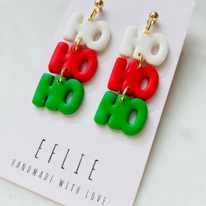 Puede incluir: Un par de pendientes colgantes con tres abalorios "HO HO HO" apilados en verde, rojo y blanco. Los pendientes están en una tarjeta blanca con el texto "EFLIE HANDMADE WITH LOVE".
