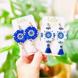 May include: Two pairs of blue and white ceramic tile earrings with gold accents and tassels. One pair has white tassels and the other has blue tassels. The earrings are displayed on white cards with the word "Efie" printed on them.