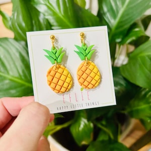 May include: A pair of gold pineapple earrings with green leaves and a yellow and orange gradient. The earrings are on a white card with the text "EFLIE HAPPY LITTLE THINGS".