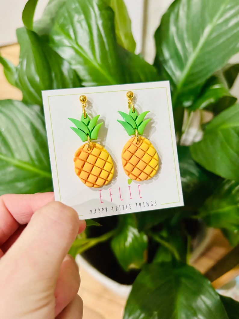 May include: A pair of gold pineapple earrings with green leaves and a yellow and orange gradient. The earrings are on a white card with the text "EFLIE HAPPY LITTLE THINGS".