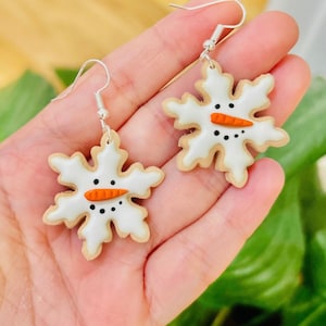 May include: A pair of snowflake-shaped earrings with a snowman face on each. The earrings are white with brown outlines and have a small orange carrot nose and black eyes.